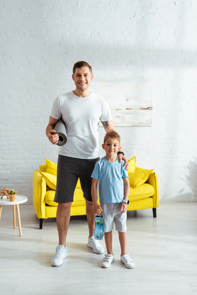 sonriente hombre con fitness mat tocando hombro de feliz hijo sosteniendo botella deportiva
 - Foto, Imagen