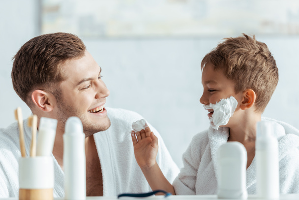 enfoque selectivo de niño feliz con espuma de afeitar en la cara mirando sonriente padre
 - Foto, imagen