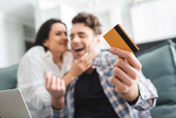 Enfoque selectivo del hombre alegre que sostiene la tarjeta de crédito cerca de la novia y del ordenador portátil en casa
  - Foto, imagen