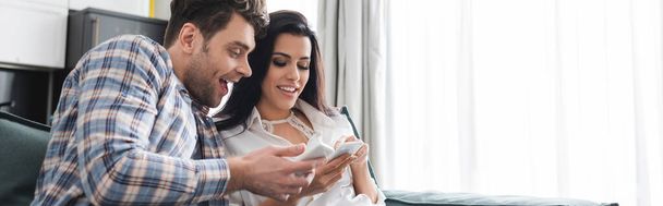 Panoramic crop of smiling couple using smartphones at home  - Foto, Imagen