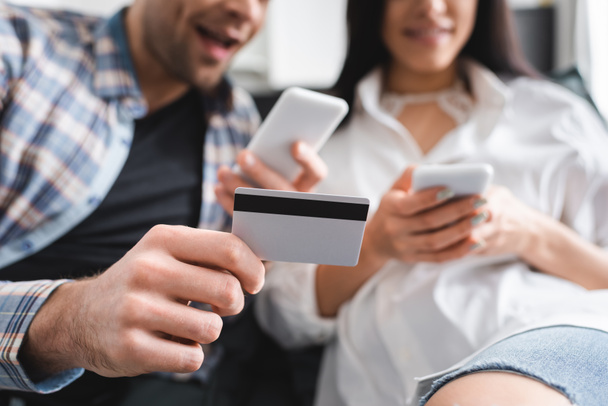 Selective focus of positive man holding credit card while using smartphone near woman  - Photo, image