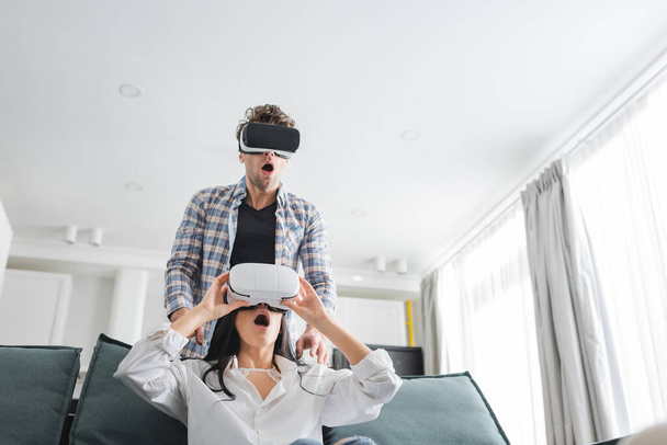 Shocked couple using virtual reality headsets in living room - Fotó, kép