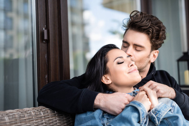 Selective focus of man kissing attractive girl on wicker sofa on terrace - Foto, imagen