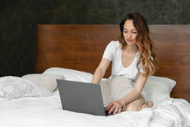 Young woman works at a laptop in the bedroom sitting on the bed. She is in pajamas and with her hair loose. Remote work. Place for an inscription. Weekend work concept at computer at home. Self-education at home with a laptop. - Photo, Image