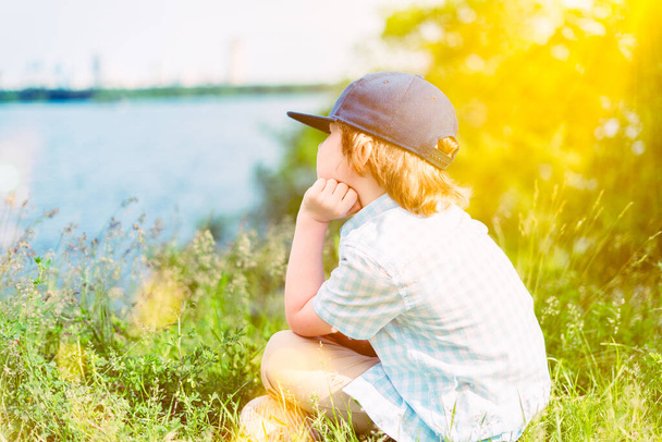 Zurück von einem kleinen Jungen, der mit Blick auf den See auf dem Gras sitzt und den Sonnenuntergang im Park beobachtet. Sommerzeit. Einsamkeit - Foto, Bild