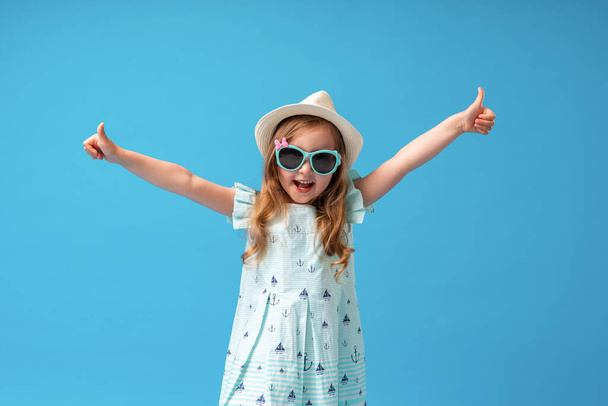 cute little girl 4 years old, in dress, hat and sunglasses, posing on blue background. child spreads his hands to side with gesture approval, will start summer vacation with pleasure and go on trip. - Photo, image