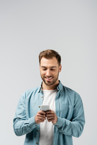usmívající se mladý muž pomocí smartphone izolované na šedé - Fotografie, Obrázek