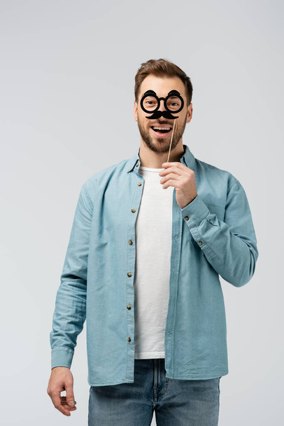 usmívající se mladý muž s falešným knírem a brýlemi na hůl izolované na šedé - Fotografie, Obrázek