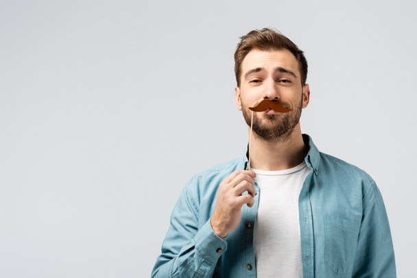 Αστείος νεαρός με ψεύτικο μουστάκι σε ξυλάκι απομονωμένος σε γκρι - Φωτογραφία, εικόνα