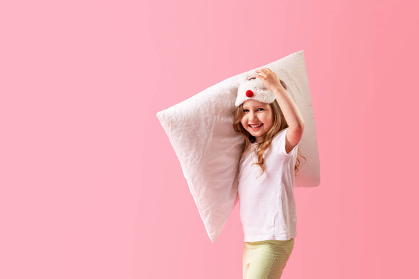 charming little girl in a sleep mask and pajamas is walking holding pillows and smiling on a pink background. The concept of healthy sleep. A copy of the space. In full growth. - Fotó, kép