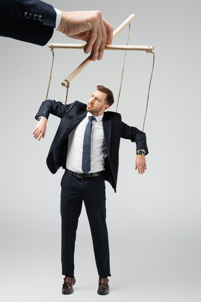 cropped view of puppeteer holding businessman marionette on strings isolated on grey - Fotoğraf, Görsel