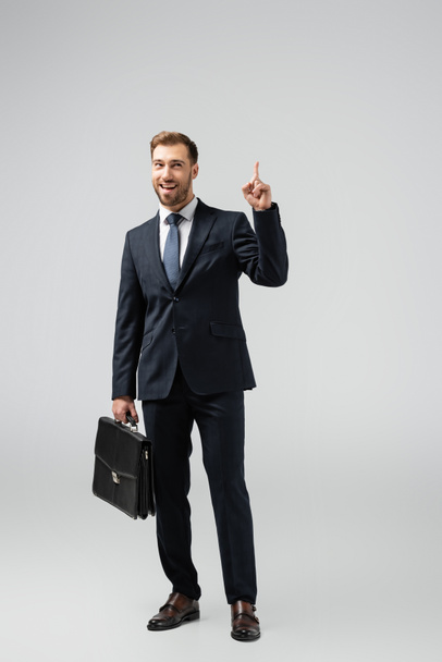 homem de negócios sorridente com mala de couro apontando para cima isolado em cinza
 - Foto, Imagem