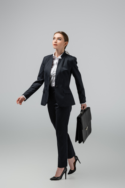 young businesswoman walking with leather suitcase isolated on grey - Zdjęcie, obraz