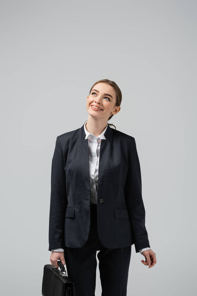 smiling young businesswoman with leather suitcase looking up isolated on grey - Фото, зображення