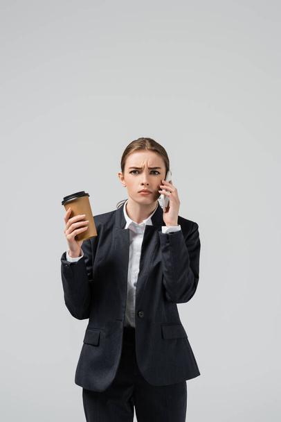 confused young businesswoman with coffee to go talking on smartphone isolated on grey - Foto, immagini