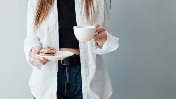 Camiseta chica de blanco y pantalones vaqueros sostiene taza de café y plato de macarrones franceses
 - Foto, Imagen