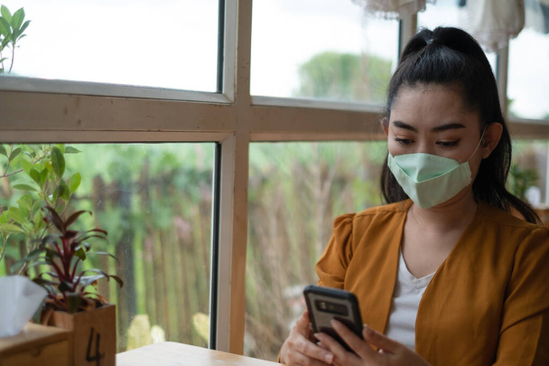 Молодая азиатка сидит и надевает медицинскую маску для защиты от вирусных инфекций дыхательных путей в кафе
 - Фото, изображение