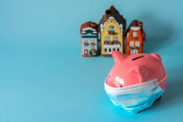 Piggy bank with medical mask and savings for real estate. Piggy bank with house models on blue background. Concept of real estate price during pandemic and quarantine. Selective focus on piggy bank - Photo, Image
