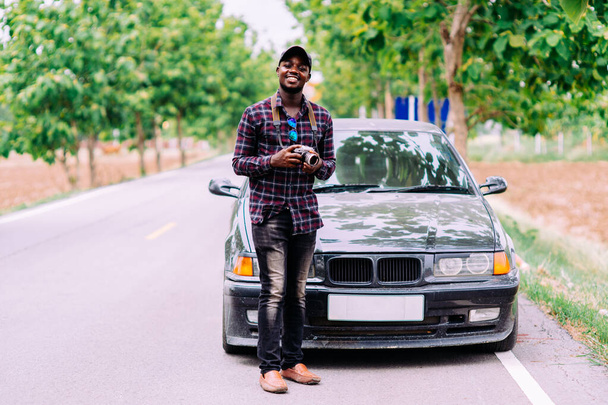 African man traveler is holding camera and standing near car freelancing on the road - Foto, imagen