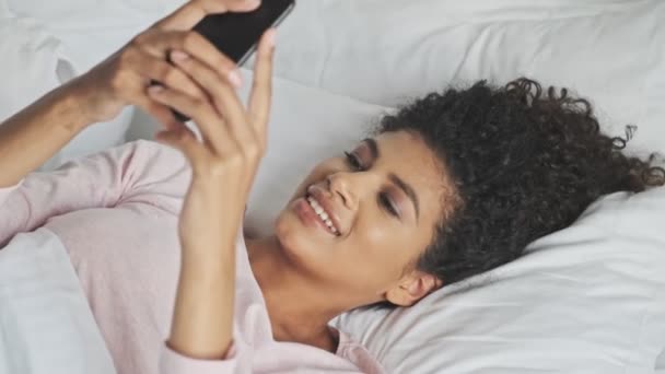 A smiling young african woman is using her smartphone lying in the bed at home - Video, Çekim