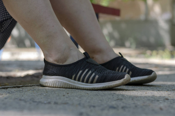 Πόδια σε αθλητικά παπούτσια μιας γυναίκας που αναπαύεται σε ένα παγκάκι. Κοντινό πλάνο με μαύρα αθλητικά. Γυναίκα 30-40 ετών. Ξεκουραστείτε κατά τη διάρκεια του καλοκαιριού περπατώντας στους δρόμους της πόλης. Επιλεκτική εστίαση. - Φωτογραφία, εικόνα