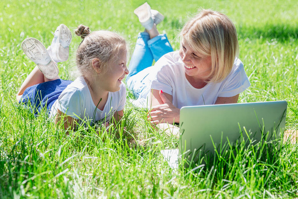 Young woman with a little daughter lie in the park with a laptop and laugh. Blogging, online training and distance learning. - Zdjęcie, obraz
