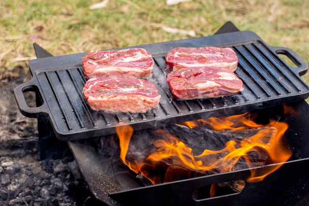 Beef steaks grilling on a cast iron plate on a camp fire. Campfire cooking. Outdoor BBQ - Photo, Image