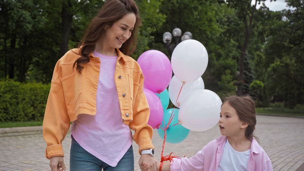 figlia parlando e tenendosi per mano con madre felice vicino palloncini nel parco - Filmati, video
