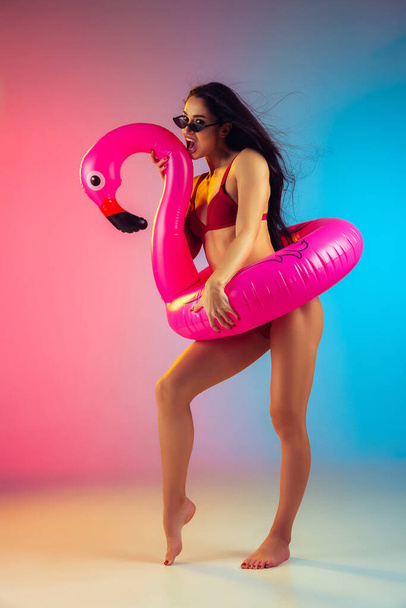 Fashion portrait of young fit and sportive woman with rubber flamingo in stylish red swimwear on gradient background. Perfect body ready for summertime. - Valokuva, kuva