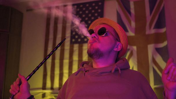 Young hookah smoker exhales up cloud of smoke in dark room with unusual neon glowing. Stylish man rests at home with American and GB flag on background - 写真・画像