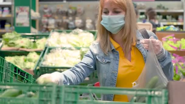 Orvosi maszkos nő és védőkesztyű választja ki és vásárolja meg az ételt a szupermarketben a covid-19 koronavírus járvány idején. Felnőtt lány felhalmoz uborkát a karantén és az önizolálás alatt. - Felvétel, videó