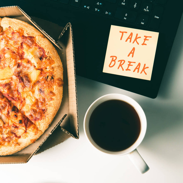 Pizza in box, cup of coffee, laptop and sticker with text TAKE A BREAK on table in office. - Fotoğraf, Görsel