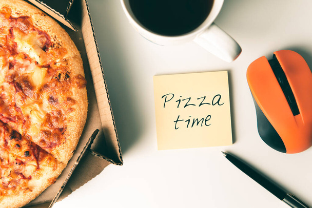Pizza in box, cup of coffee, laptop, computer mouse, pen and empty sticker on table in office. - Foto, imagen