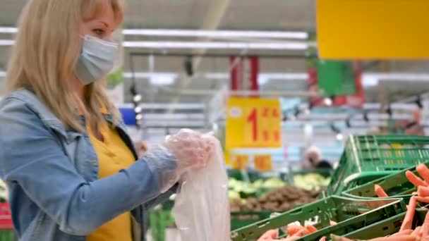 Tıbbi maske takan ve koruyucu eldivenli bir kadın kovid 19 Coronavirus salgını sırasında süpermarketten yiyecek seçip alıyor. Yetişkin kız karantina ve kendini izole etme süresince havuç stokluyor.. - Video, Çekim