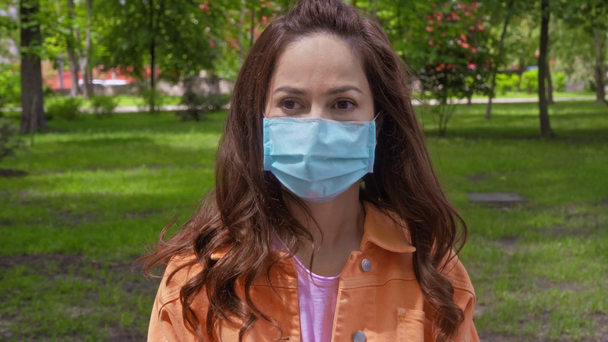 γυναίκα με μπλε ιατρική μάσκα στέκεται στο πάρκο  - Πλάνα, βίντεο