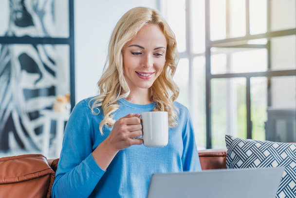 Young woman working on laptop computer while sitting on sofa at home with morning cup of coffee - Photo, image