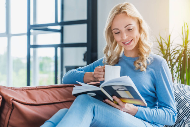 Young woman at home sitting on modern sofa relaxing in living room while reading book and drinking coffee - Foto, afbeelding