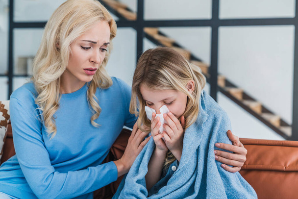 Portrait of caucasian mother hugging her ill daughter while sneezing in tissue under blanket at home - Фото, изображение