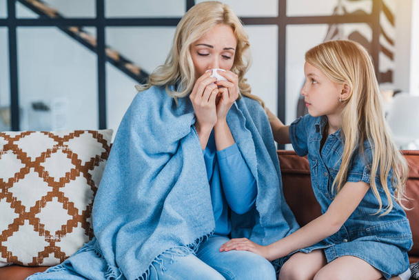Little cute girl sitting with her mother on sofa while mother sneezing in tissue - 写真・画像