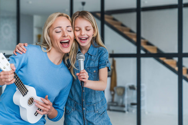 Beautiful mother and her cute little daughter with microphone singing together and playing ukulele - Fotoğraf, Görsel