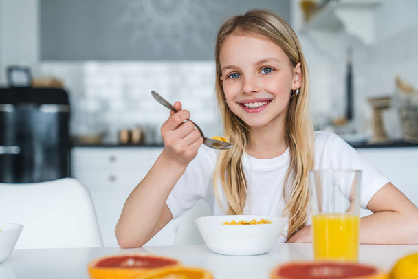 Cute little girl looking at camera while eating cereal balls with juice for her breakfast - Фото, изображение