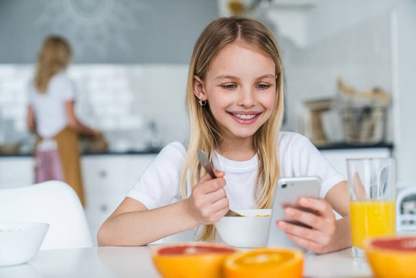 Little girl having breakfast while using phone at home kitchen - Foto, Imagem