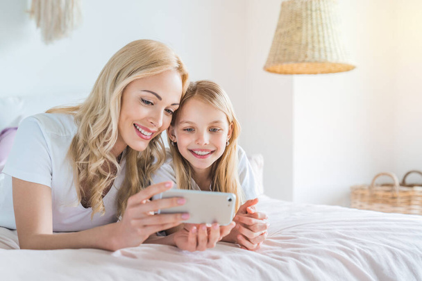Happy young woman lying on bed with smiling kid daughter making selfie photo on smartphone or using it - Foto, Imagen