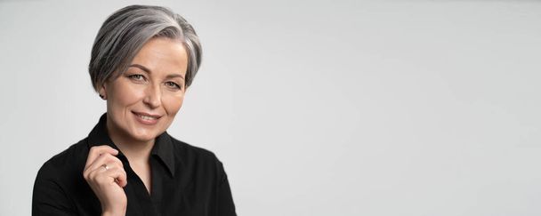Pretty mature woman smiles with hand near face. Charming gray-haired lady in black blouse on white background. Copy spase for text. Horizontal blank or template for ad banner - Fotoğraf, Görsel