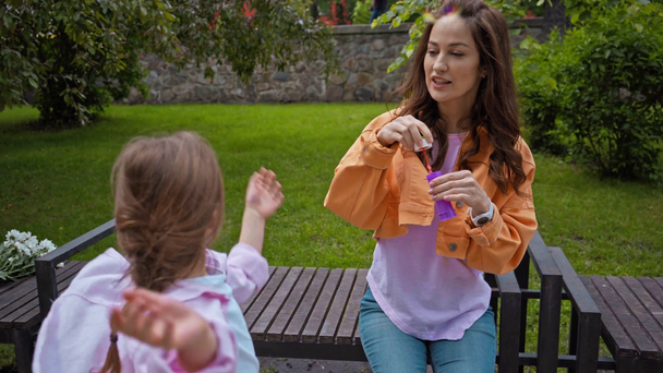 attractive mother blowing soap bubbles near cute daughter in park  - Imágenes, Vídeo