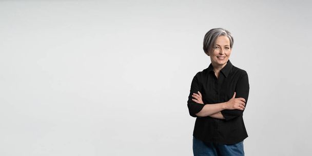 Confident mature woman smiling arms crossed. Gray haired businesswoman in black shirt on white background. Horizontal template for ad banner with copy spase for text - Фото, изображение