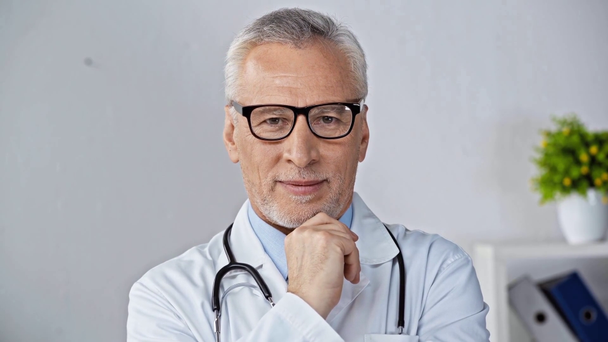 mature doctor in glasses smiling, touching chin and waving hand - Footage, Video