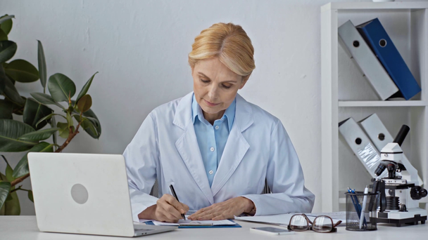 зрелый и привлекательный врач с помощью ноутбука рядом с документами в клинике
 - Кадры, видео