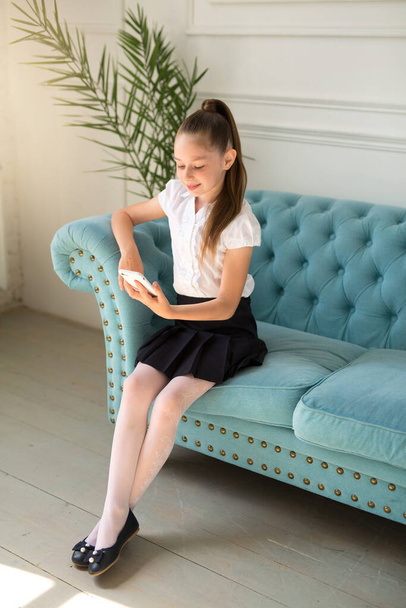  beautiful little teenager girl in a white shirt and skirt sits on a sofa with a phone - Foto, immagini