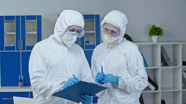 científico en traje de materiales peligrosos hablando con su compañero de trabajo y escribiendo en el portapapeles
  - Metraje, vídeo